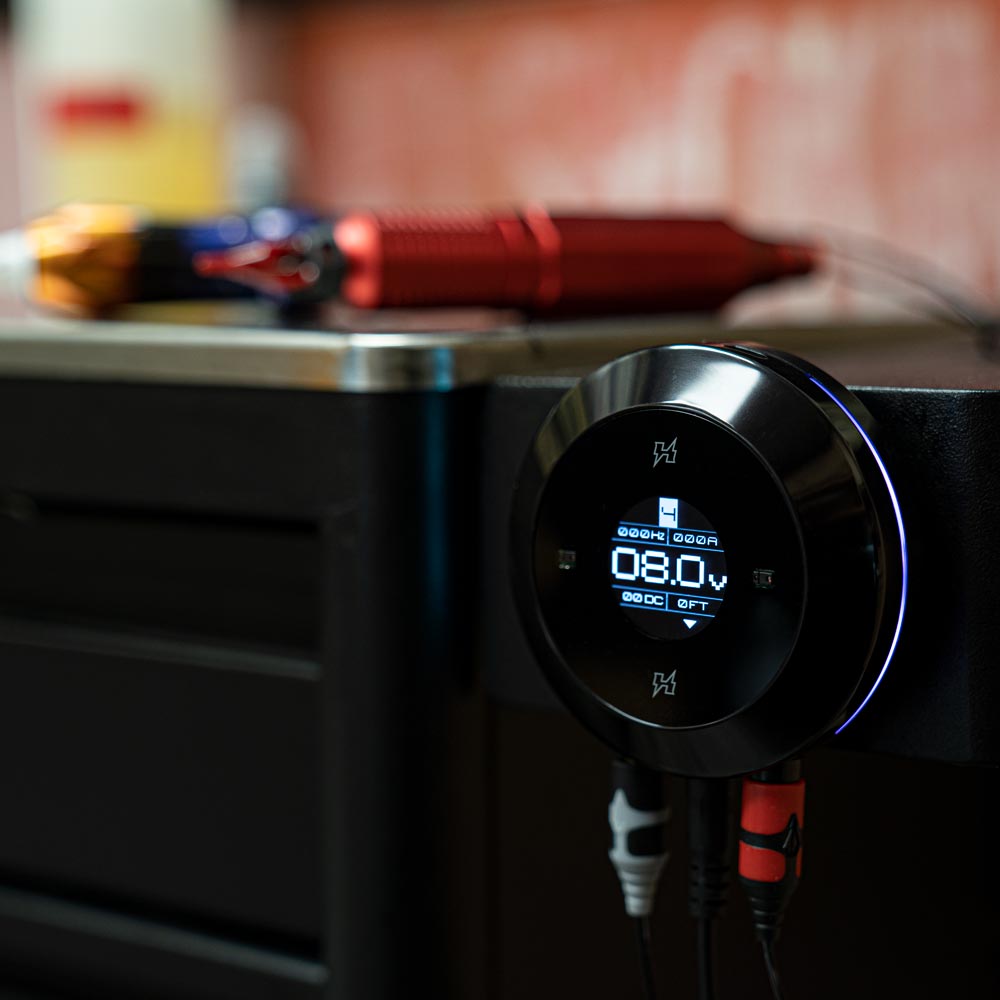 FK Irons Hover Touchless Tattoo Power Supply in focus with machines in background